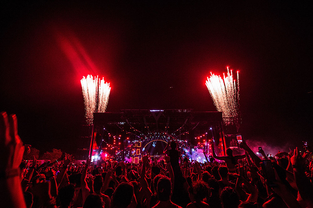 Concert crowd at night facing the stage with a reddish lights and fireworks on either side